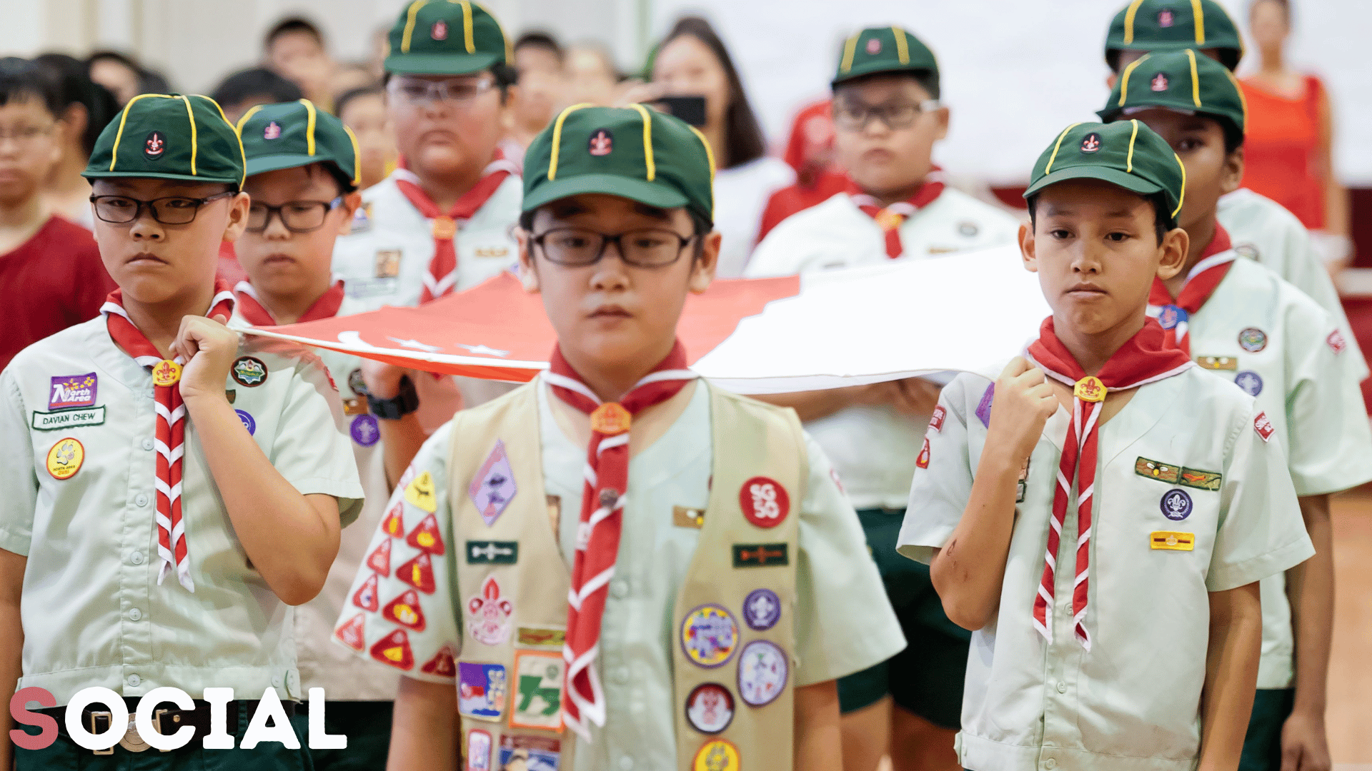 Boy scouts in green caps, white shirts, red neckerchiefs, and merit badge sashes carry a folded flag indoors.